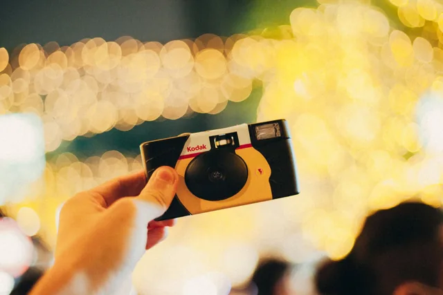 A hand holding a disposable camera against a background of blurred fairy lights