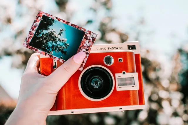 A hand holding a red instant camera with a freshly developed photo